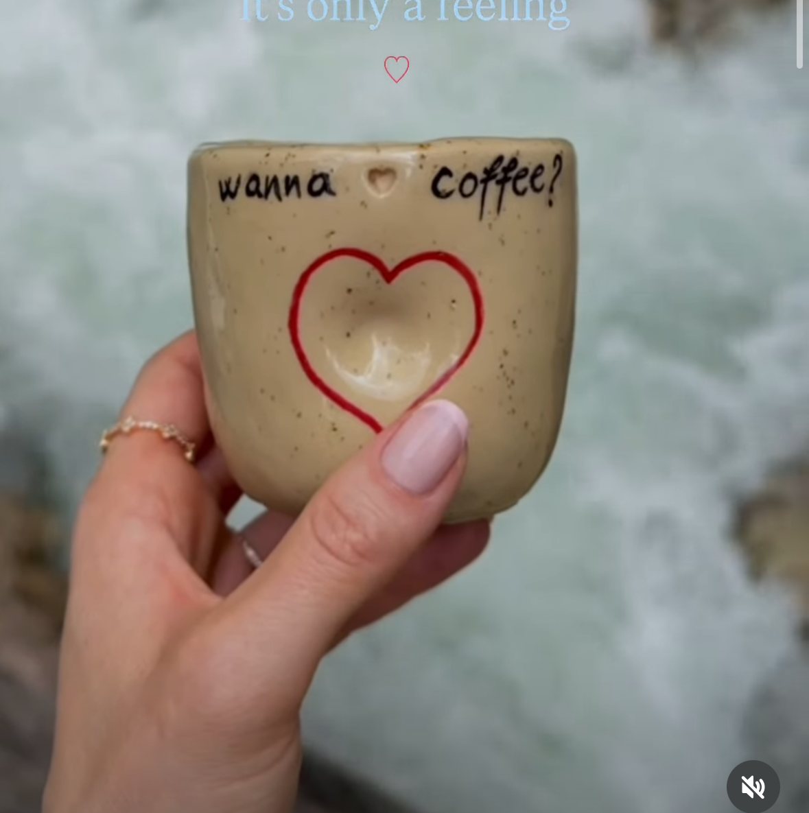 Handmade speckled ceramic mug hand-lettered with 'wanna coffee?' and a red heart, held over a mountain river